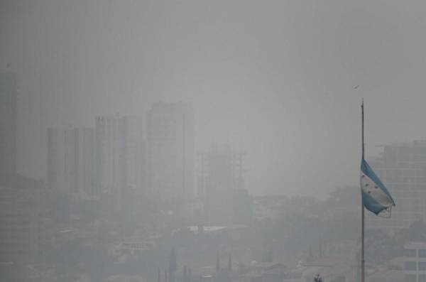 <i>Una bandera nacional hondureña ondea en medio de una densa capa de humo que cubre Tegucigalpa el 6 de mayo de 2024. La capital hondureña enfrenta problemas de contaminación debido a la gran cantidad de vehículos e incendios forestales que ocurren en la región. FOTO Orlando SIERRA/AFP</i>