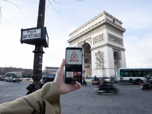 <i>Una mujer sostiene su teléfono móvil con la aplicación de alojamiento en casas de corta y larga duración Airbnb en la pantalla mientras se encuentra en la Place Charles de Gaule, mirando el Arco de Triunfo en el centro de París el 2 de febrero de 2024. FOTO MIGUEL MEDINA / AFP</i>